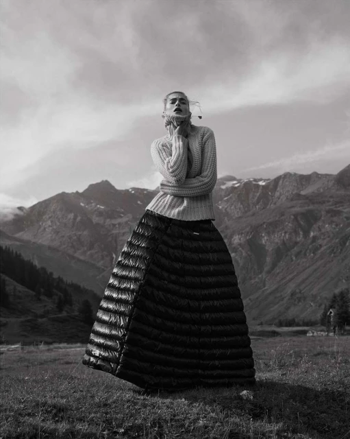 Monochrome fashion shot of a woman in a sweater and quilted skirt, posing in a field with mountains and a cloudy sky in the background.