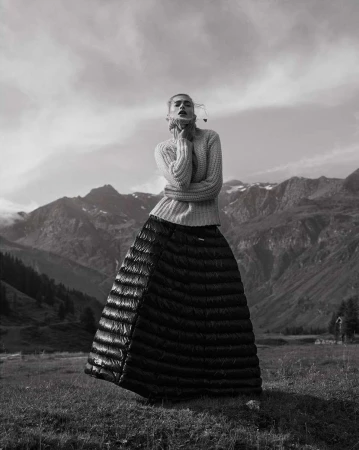 Monochrome fashion shot of a woman in a sweater and quilted skirt, posing in a field with mountains and a cloudy sky in the background.