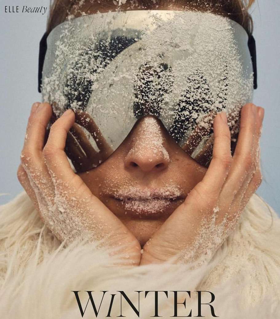Close-up of a woman's face covered in snow, wearing ski goggles and a white fur coat against a light blue background.