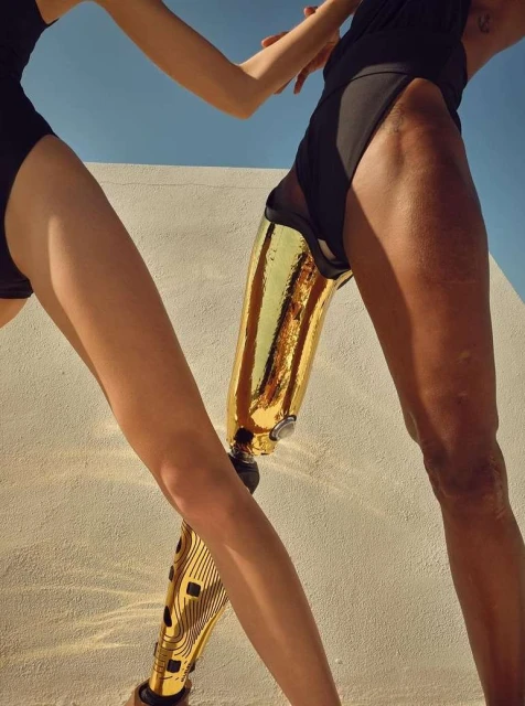 Two women pose in black swimsuits against a white wall with a blue sky, one with a gold prosthetic leg, for a fashion-related photograph.