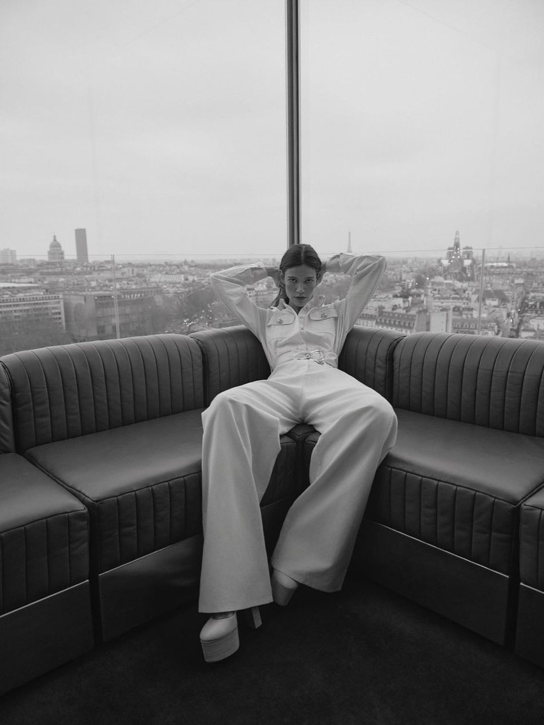 Black and white fashion shot of a woman in a jumpsuit sitting on a couch with a cityscape visible through a large window.