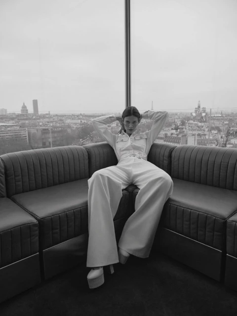 Black and white fashion shot of a woman in a jumpsuit sitting on a couch with a cityscape visible through a large window.
