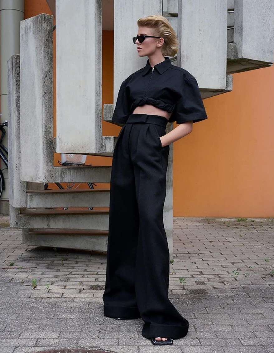 Model in black sunglasses and black two-piece outfit standing outdoors near concrete architectural structure