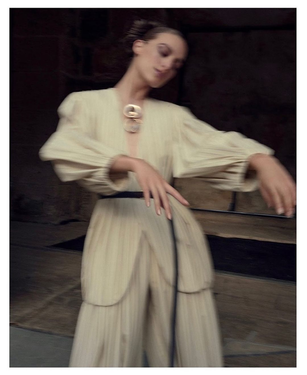 Model in cream pleated tunic and trousers with a black belt and statement necklace posing outdoors in motion blur.