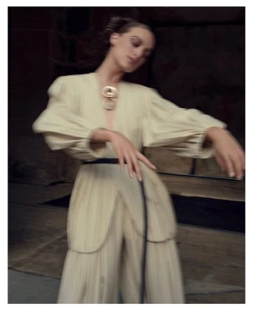 Model in cream pleated tunic and trousers with a black belt and statement necklace posing outdoors in motion blur.
