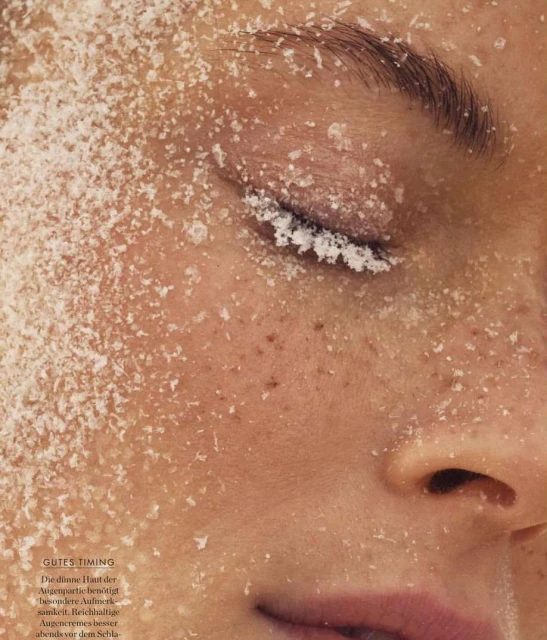 Close-up of a woman's face with eyes closed, face and hair covered in white flakes, possibly snow or dandruff. Beauty shot with skin texture.