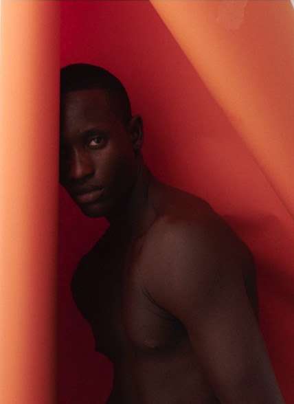 Headshot of a dark-skinned man leaning against a textured orange wall, highlighting his face and bare chest in a minimalist setting.