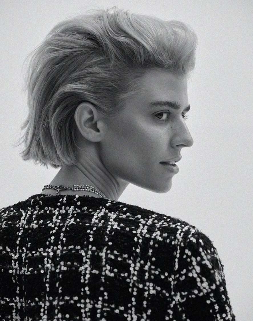 Woman in black and white tweed jacket with blond bob hairstyle in a studio headshot, facing away from camera, looking to the right.