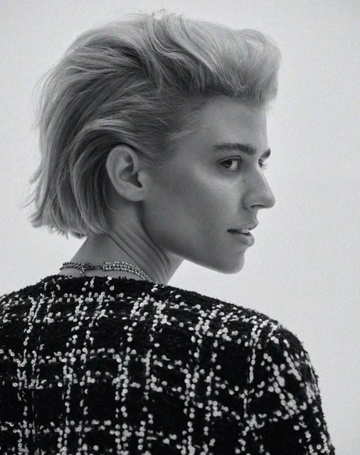 Woman in black and white tweed jacket with blond bob hairstyle in a studio headshot, facing away from camera, looking to the right.