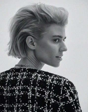 Woman in black and white tweed jacket with blond bob hairstyle in a studio headshot, facing away from camera, looking to the right.