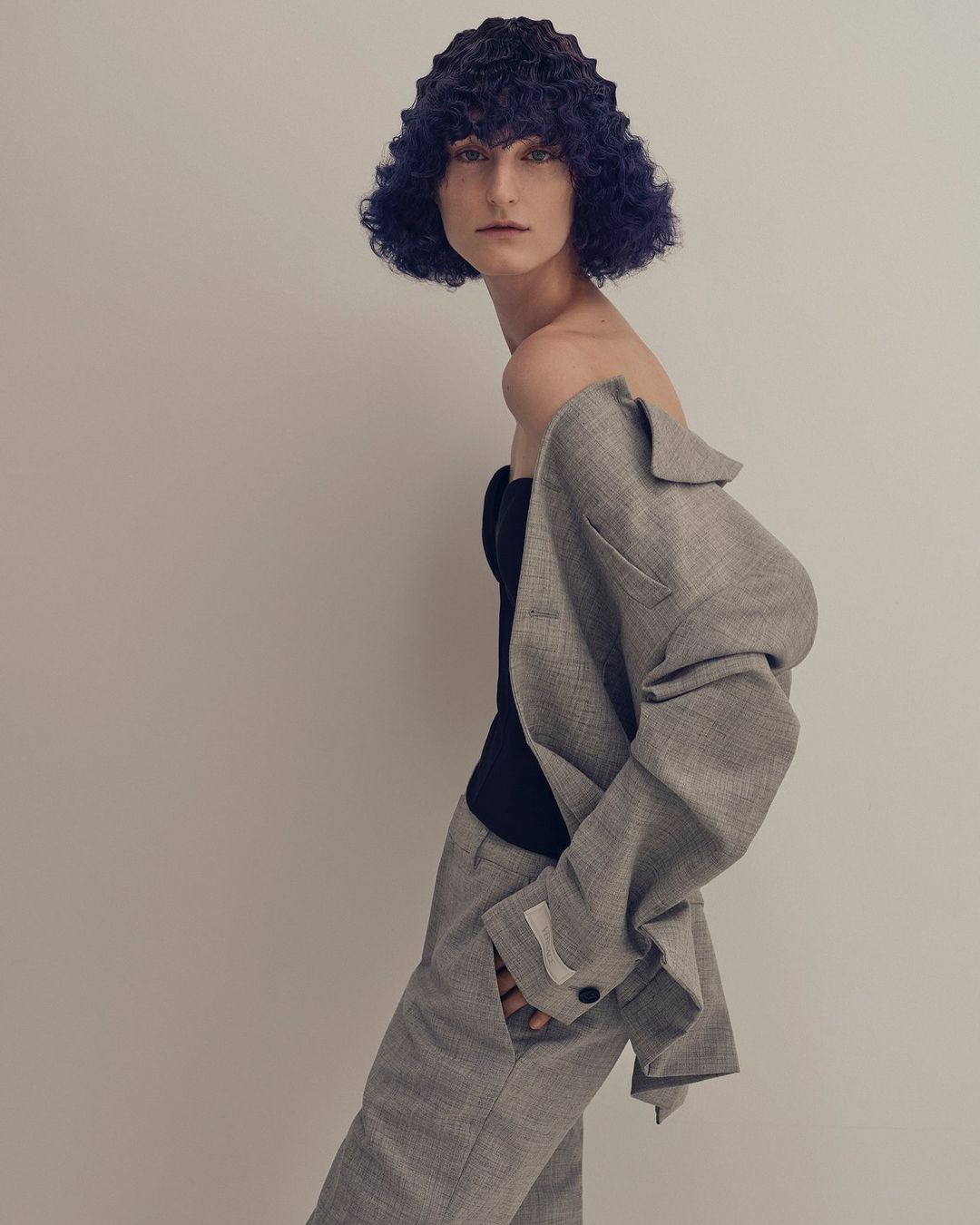 Portrait of a model in a gray suit with blue textured hair, posing in front of a neutral background, fashion photography.