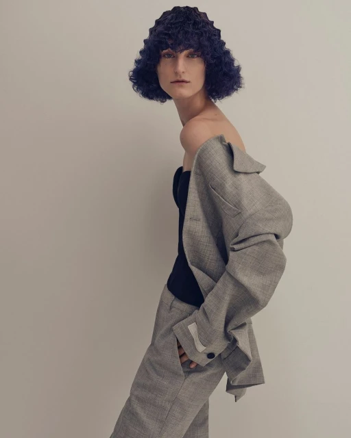 Portrait of a model in a gray suit with blue textured hair, posing in front of a neutral background, fashion photography.
