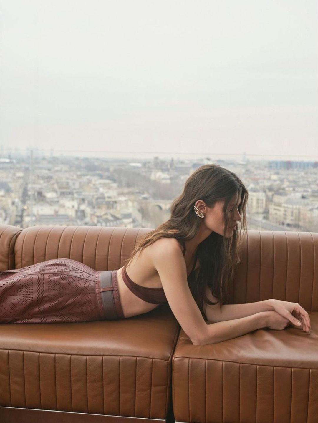Adult woman lying on a brown leather sofa in front of a cityscape, wearing a brown two-piece outfit and statement earrings.