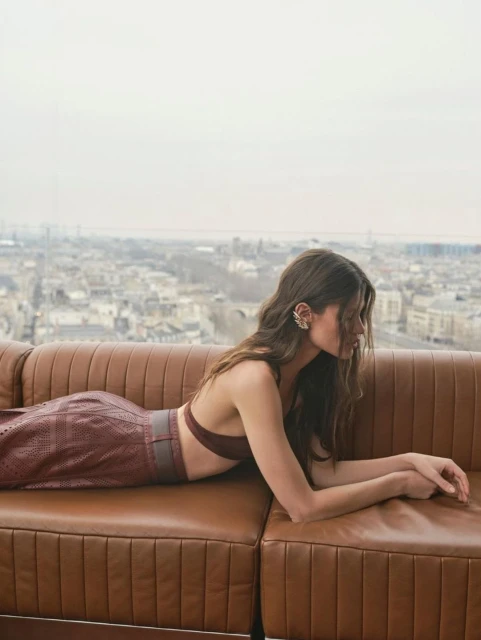 Adult woman lying on a brown leather sofa in front of a cityscape, wearing a brown two-piece outfit and statement earrings.