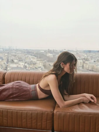 Adult woman lying on a brown leather sofa in front of a cityscape, wearing a brown two-piece outfit and statement earrings.