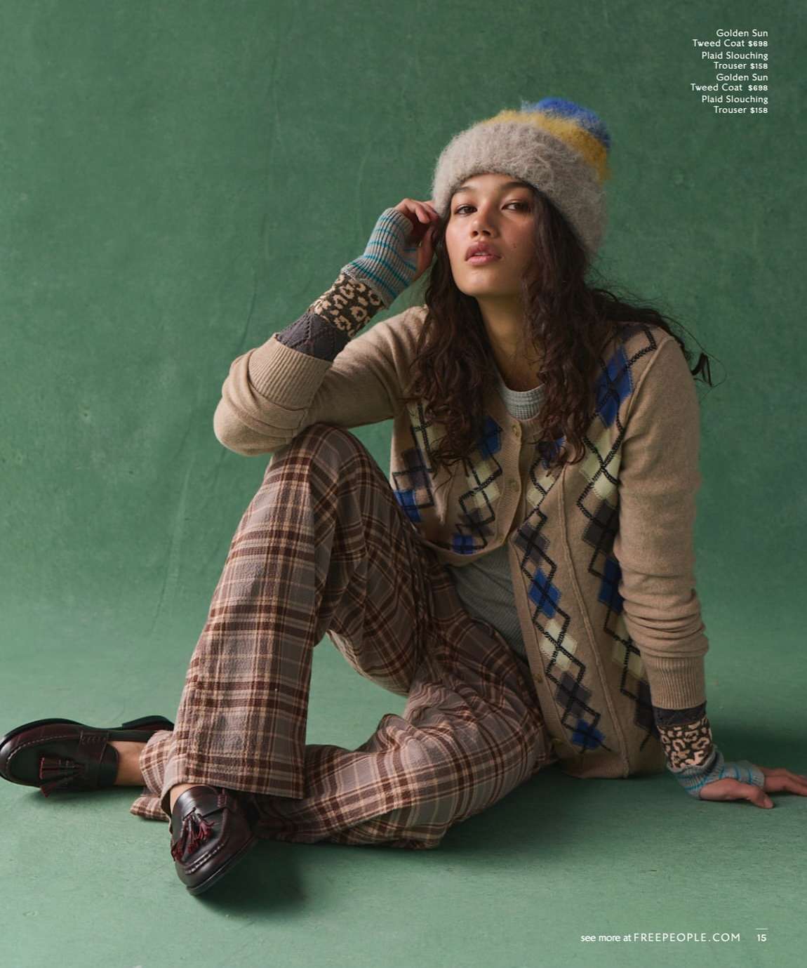 A woman poses in fashionable knit hat, cardigan, plaid pants, and loafers against a green backdrop in a studio fashion shot.