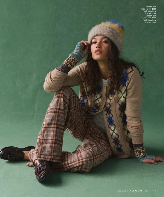 A woman poses in fashionable knit hat, cardigan, plaid pants, and loafers against a green backdrop in a studio fashion shot.