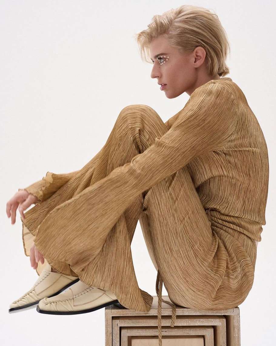 Woman in gold pleated outfit sits on wooden box in studio. Blonde short hair and embellished eyes. Fashion portrait on white background.