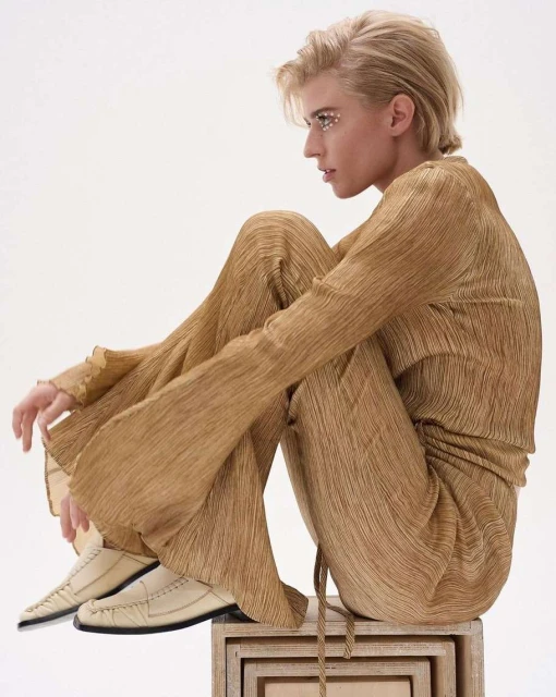 Woman in gold pleated outfit sits on wooden box in studio. Blonde short hair and embellished eyes. Fashion portrait on white background.