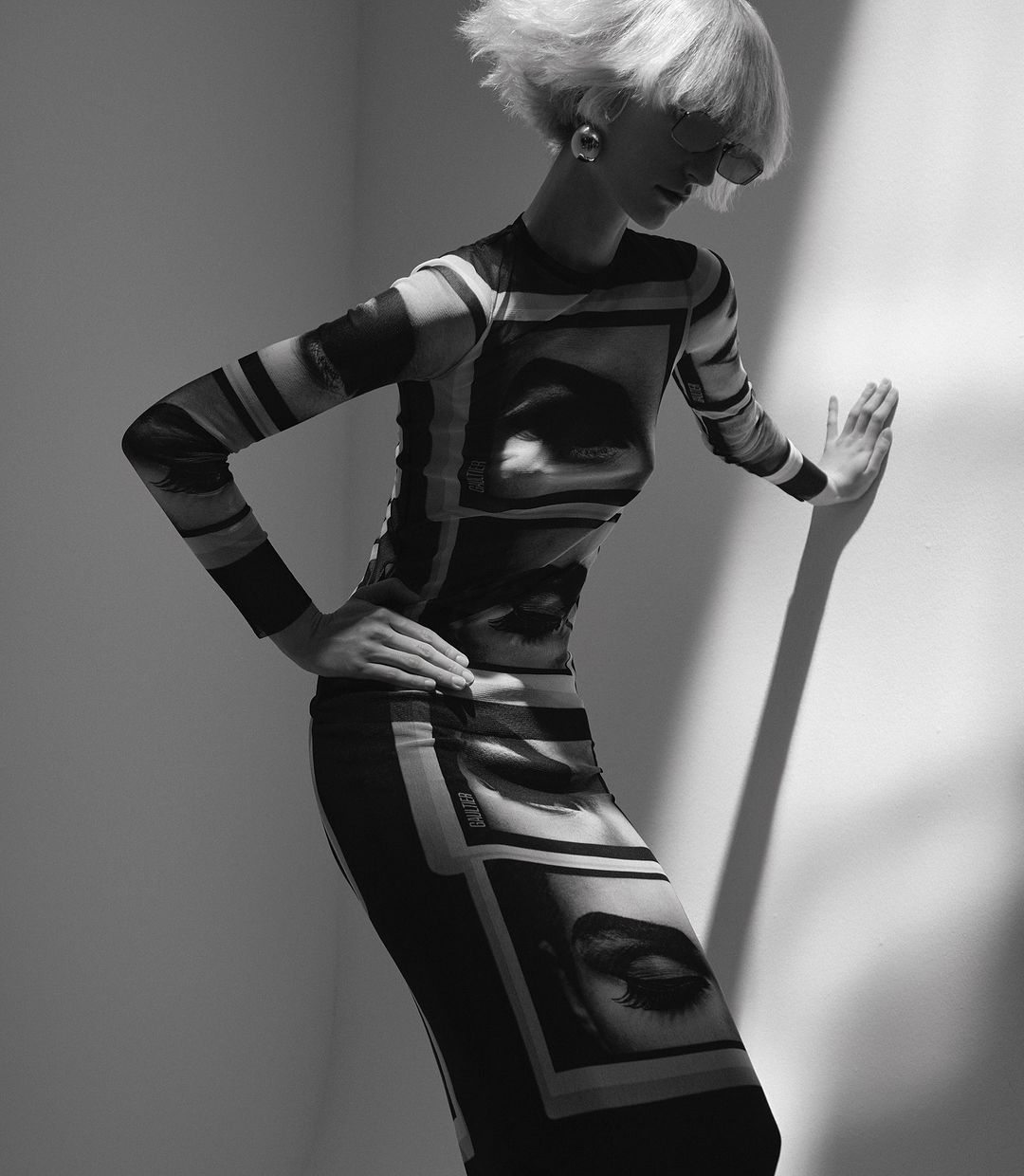 Model with blonde bob wearing a long-sleeved graphic print dress leaning against a white studio wall in black and white.