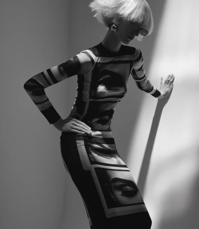 Model with blonde bob wearing a long-sleeved graphic print dress leaning against a white studio wall in black and white.