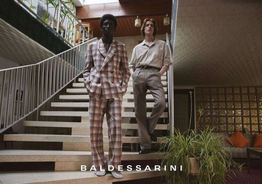 Two men model tailored menswear on a modern staircase. One wears a plaid suit and the other a shirt and pants. Plants and architecture visible.