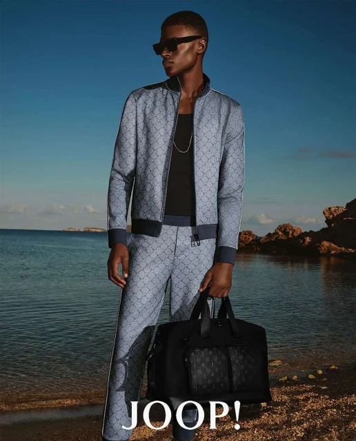 A black male model stands confidently on a beach wearing a matching patterned jacket and pants, holding a black bag with the JOOP! logo.