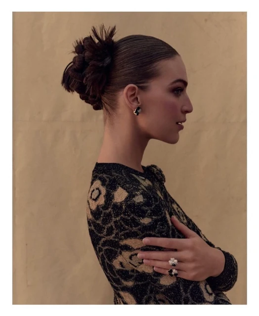Glamour shot: Young woman with braided updo, black and tan dress, floral rings, and statement earrings poses against a tan backdrop.