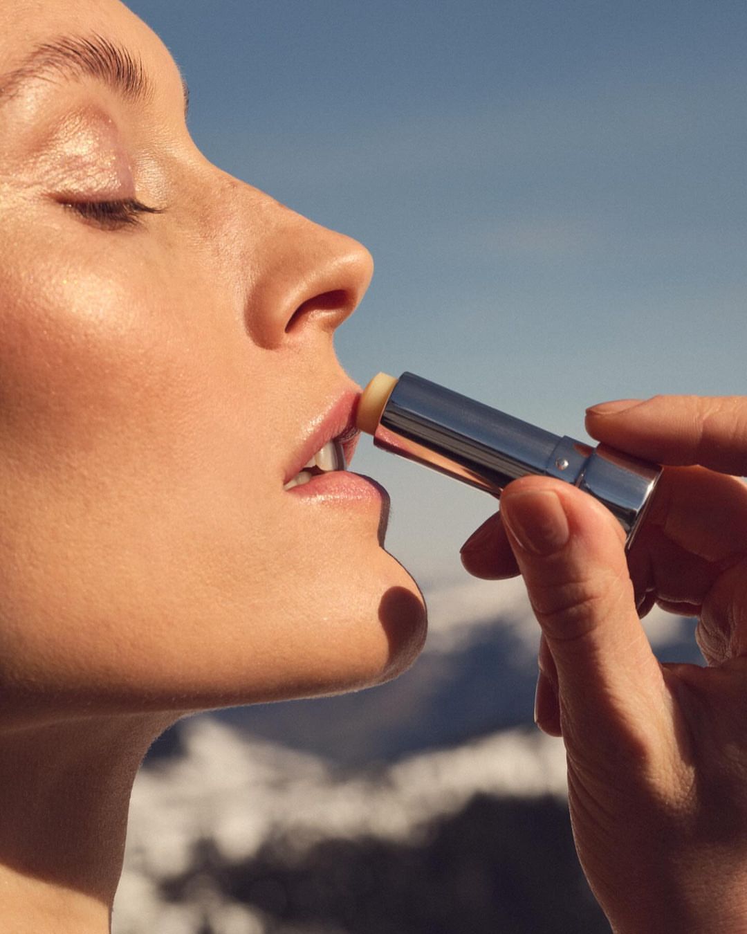 A fair-skinned adult woman applies lip balm in a close-up shot against a blue sky and blurred mountain backdrop on a sunny day.