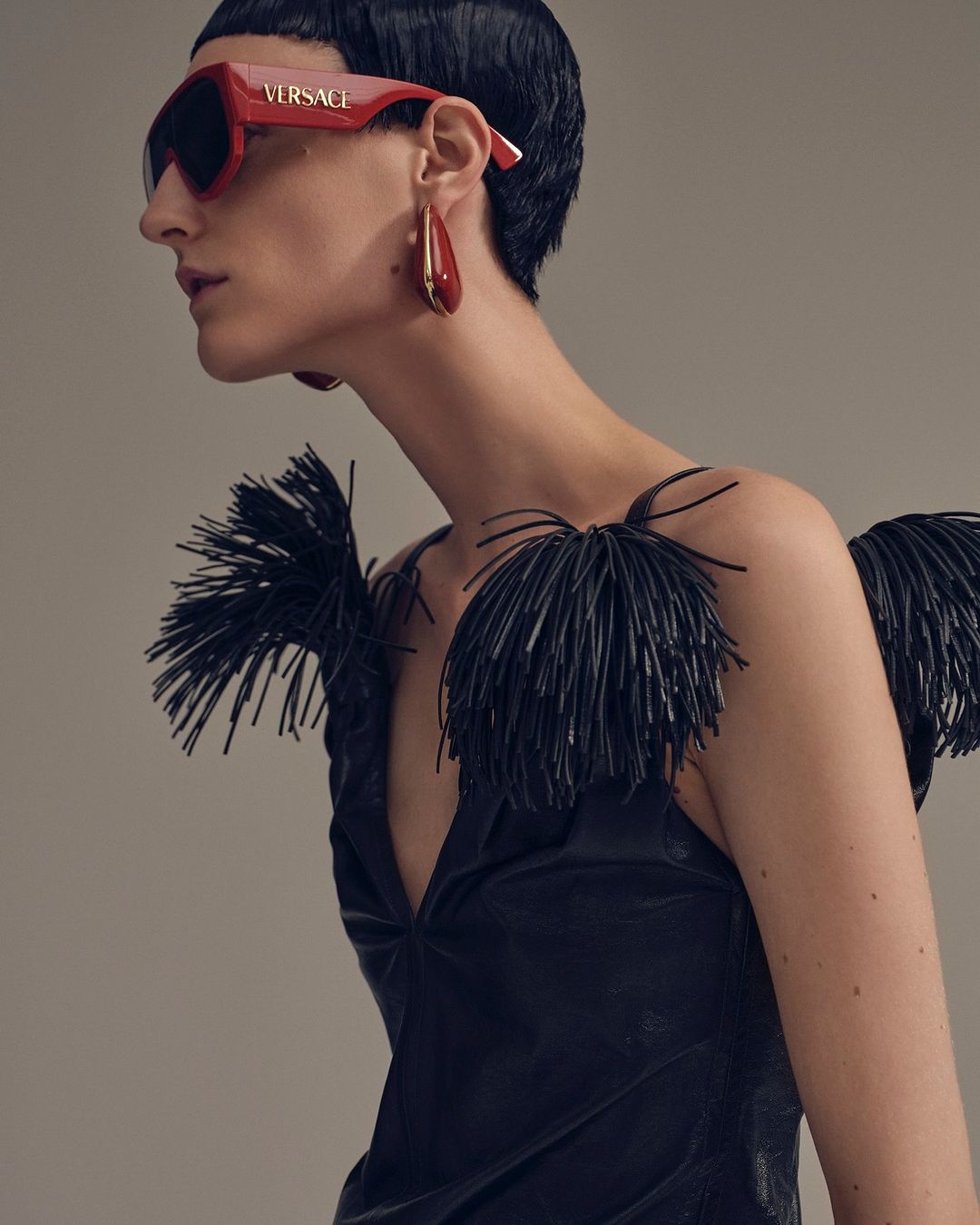 Fashionable adult female with short dark hair wearing a black dress with black feathered details, red Versace sunglasses, and red earrings.