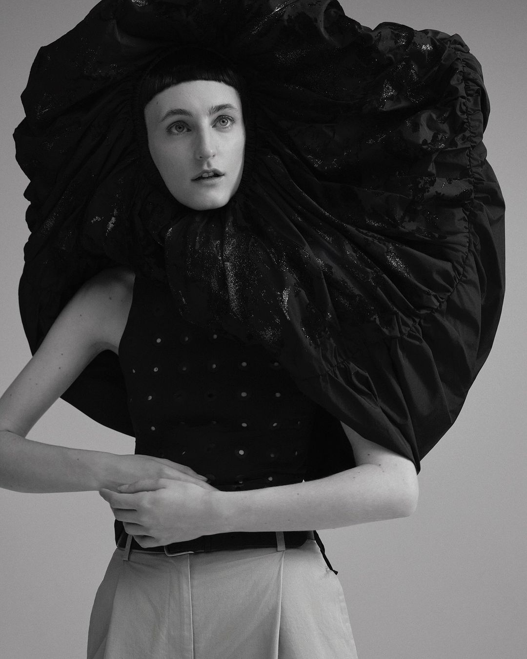 Black-and-white portrait of a woman in a sleeveless top, light pants, and large, theatrical black headpiece against a neutral background.