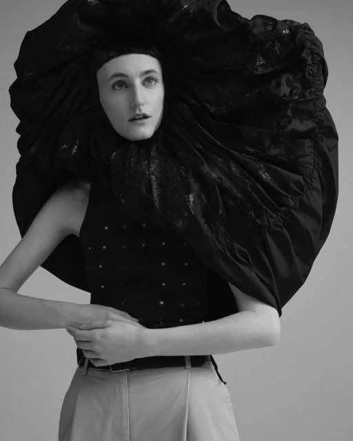 Black-and-white portrait of a woman in a sleeveless top, light pants, and large, theatrical black headpiece against a neutral background.