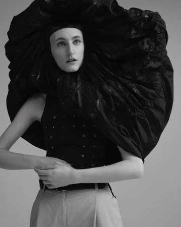 Black-and-white portrait of a woman in a sleeveless top, light pants, and large, theatrical black headpiece against a neutral background.