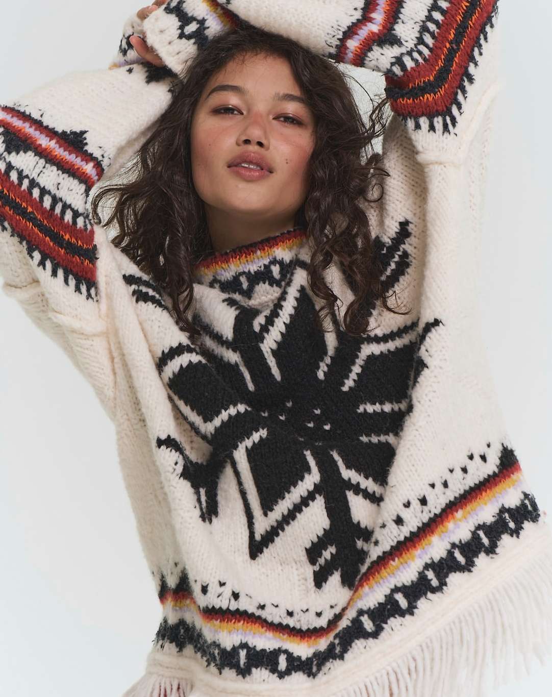 Model with dark curly hair wearing an oversized cream patterned knit sweater with fringe details against a white background