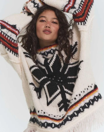 Model with dark curly hair wearing an oversized cream patterned knit sweater with fringe details against a white background