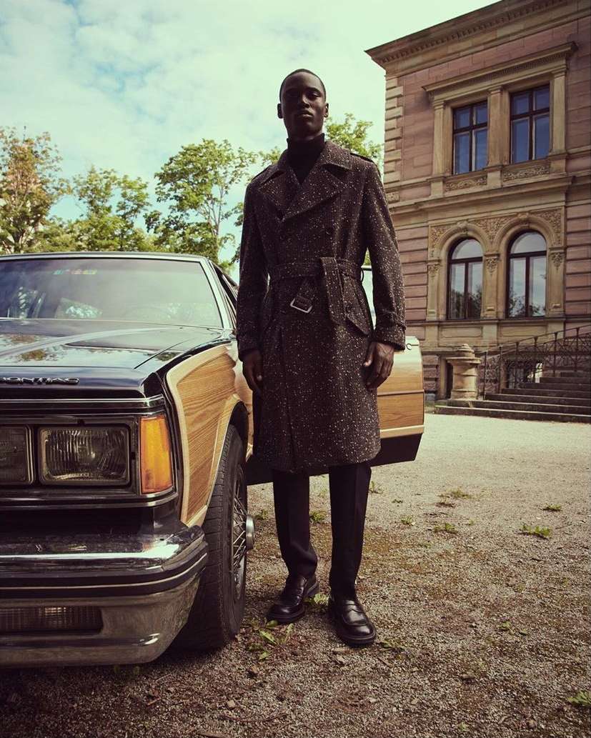 An African-American man in a tweed coat stands next to a classic woody station wagon car in front of a building on a sunny day.