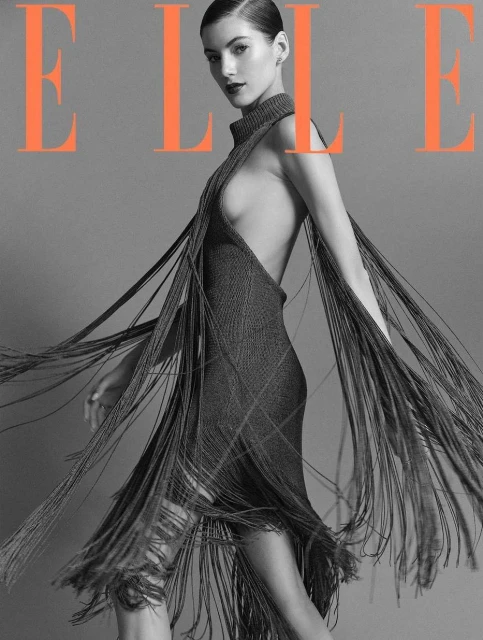 A woman poses on a fashion magazine cover in a gray fringed dress and dark lipstick against a gray background with orange magazine text.