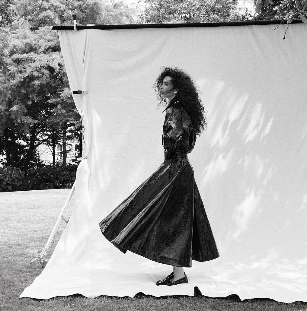Black and white portrait of a woman with long curly hair wearing a dark belted trench coat standing in front of an outdoor photography backdrop