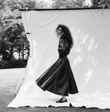 Black and white portrait of a woman with long curly hair wearing a dark belted trench coat standing in front of an outdoor photography backdrop