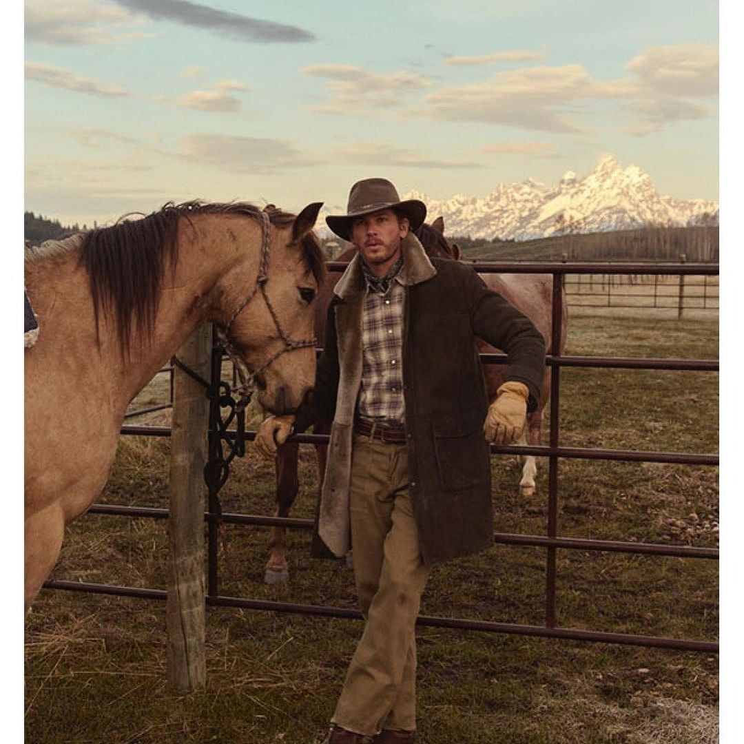A man in a cowboy hat and coat stands beside a light-brown horse in a rural landscape with mountains in the distance. 
