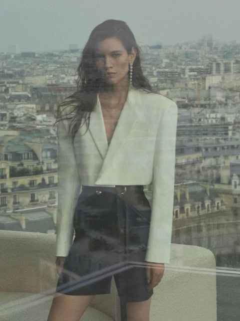 A woman in a white jacket and black shorts stands against a city backdrop with architectural features in a fashion editorial style.