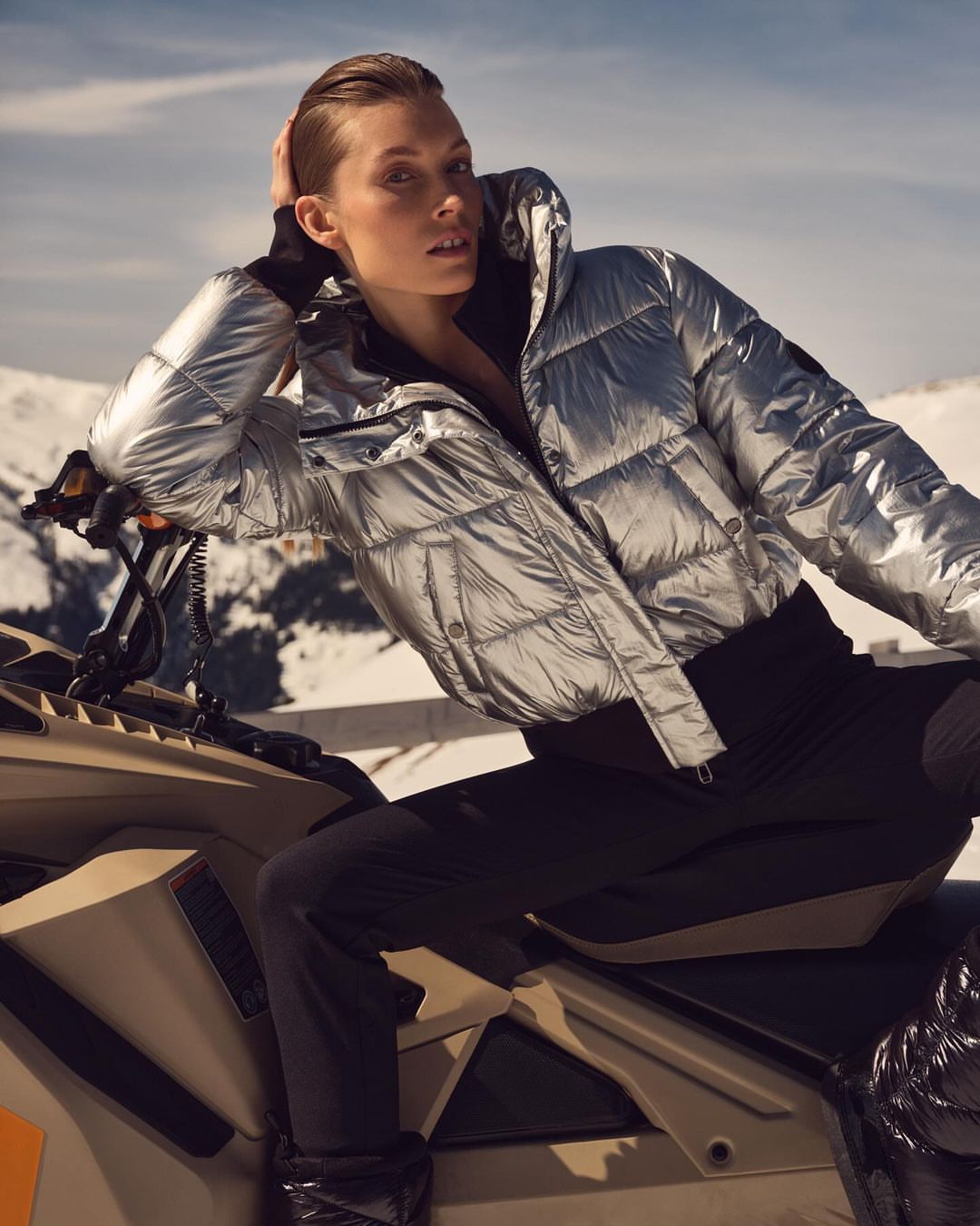 Adult woman in a silver puffer jacket poses while sitting on a snowmobile, set against a backdrop of snow-covered mountains and clear sky.