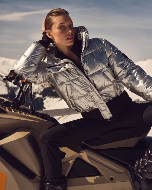 Adult woman in a silver puffer jacket poses while sitting on a snowmobile, set against a backdrop of snow-covered mountains and clear sky.