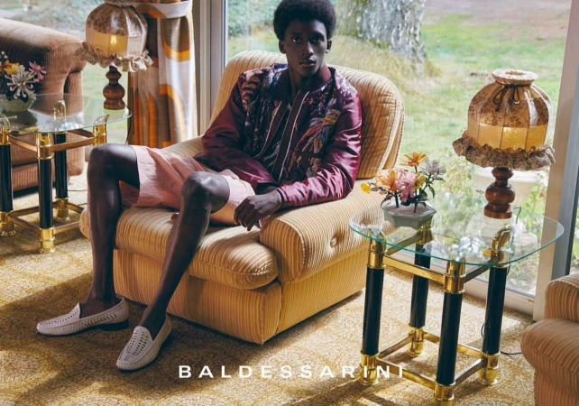 A Black man seated in a yellow armchair wearing a maroon patterned jacket, pink shorts, and white woven loafers.