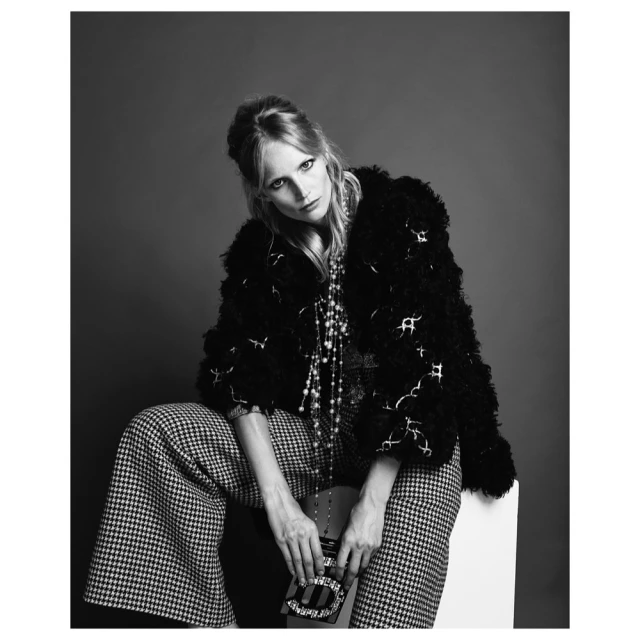 Black and white portrait of a young woman with fair skin, wearing a patterned jacket and wide-legged pants, holding a clutch purse.