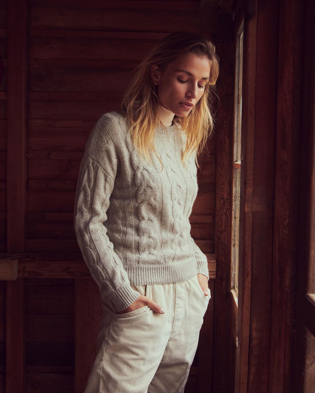A woman with blond hair wears a cable-knit sweater and cream pants in a softly lit interior with brown wood walls and a window.