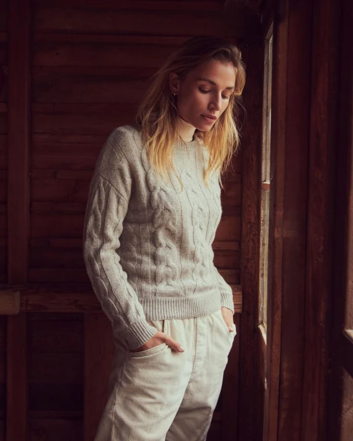 A woman with blond hair wears a cable-knit sweater and cream pants in a softly lit interior with brown wood walls and a window.