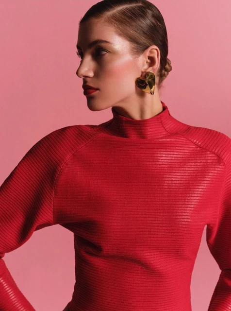 Woman in red turtleneck poses in profile with gold heart earrings against pink backdrop in a studio shot for fashion editorial.