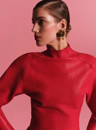 Woman in red turtleneck poses in profile with gold heart earrings against pink backdrop in a studio shot for fashion editorial.