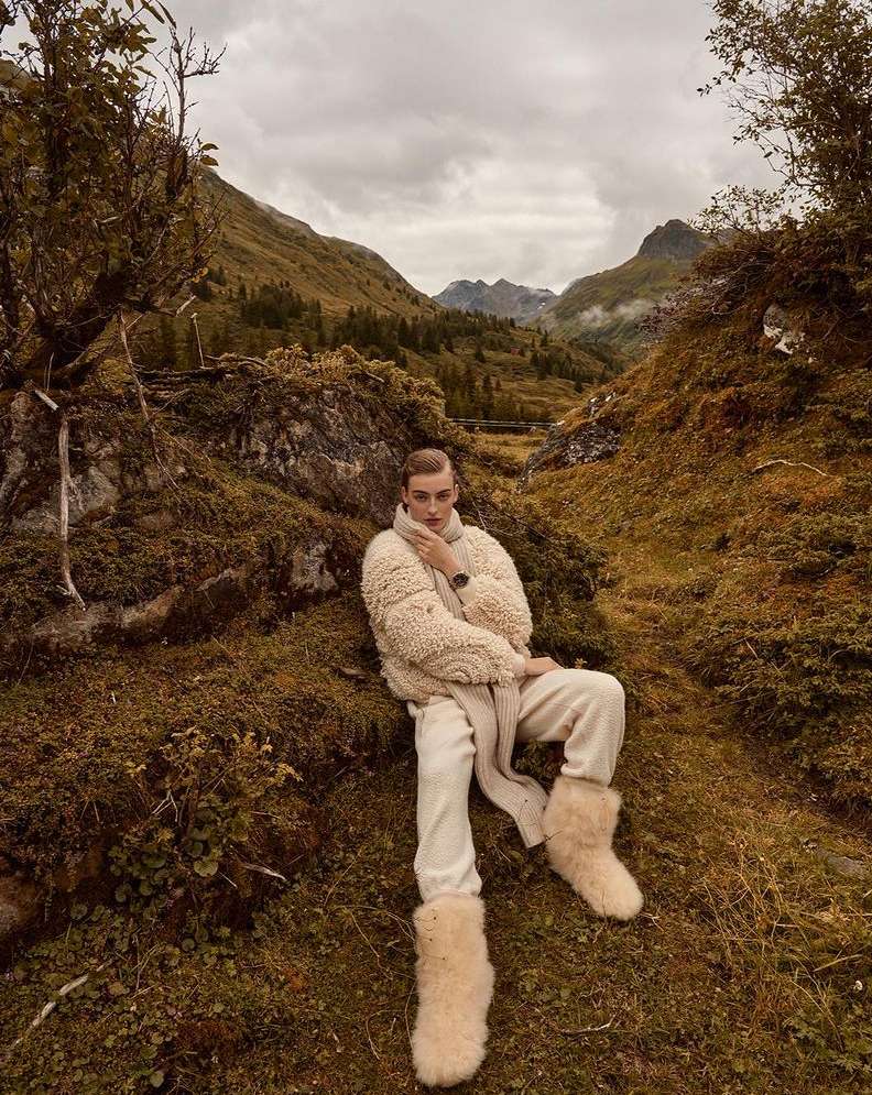 A young adult male sits in a mountainous landscape wearing a beige wool coat, trousers, scarf, and fur boots in an outdoor fashion shot.